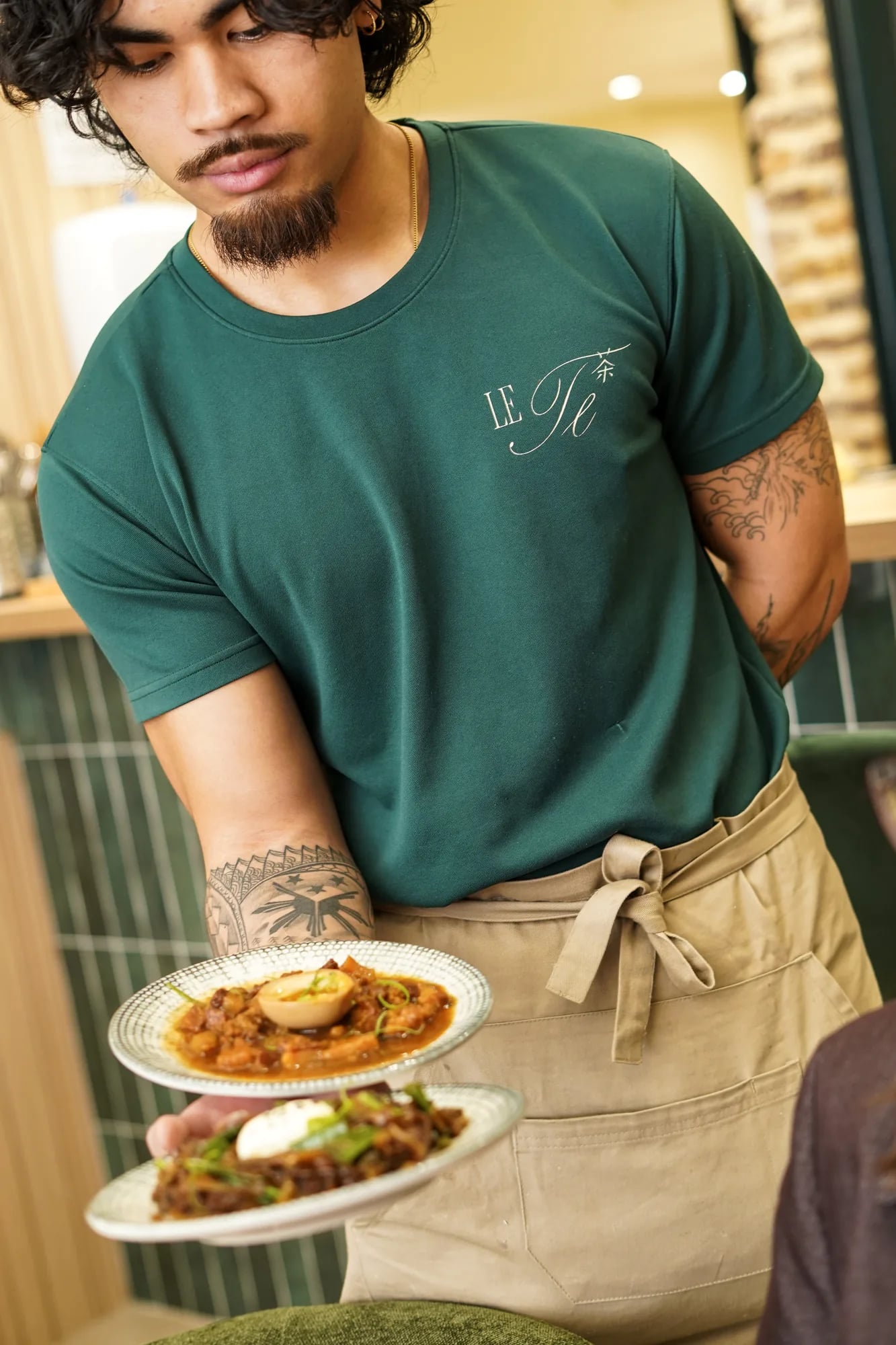 Serveur en t-shirt LE TE vert sert deux assiettes, tatouages bras